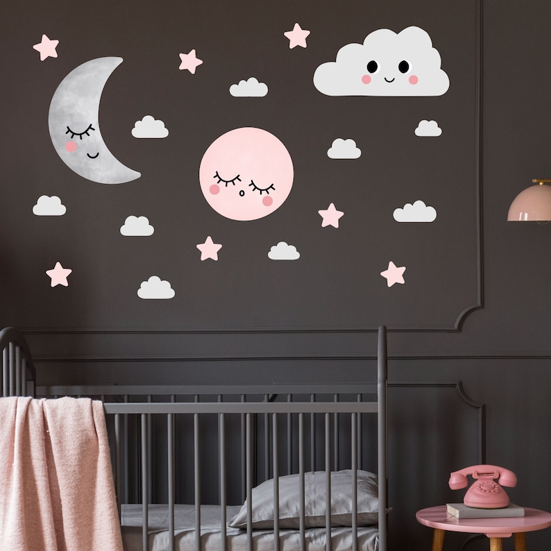 May include: A nursery wall decal set with a pink and gray color scheme. The set includes a crescent moon, a sun, a cloud, and stars. The moon, sun, and cloud have cute faces.