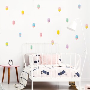 May include: A white metal bed frame with a pink and white patterned duvet and a white pillow with a whale design. The wall behind the bed is decorated with colorful popsicle stickers.