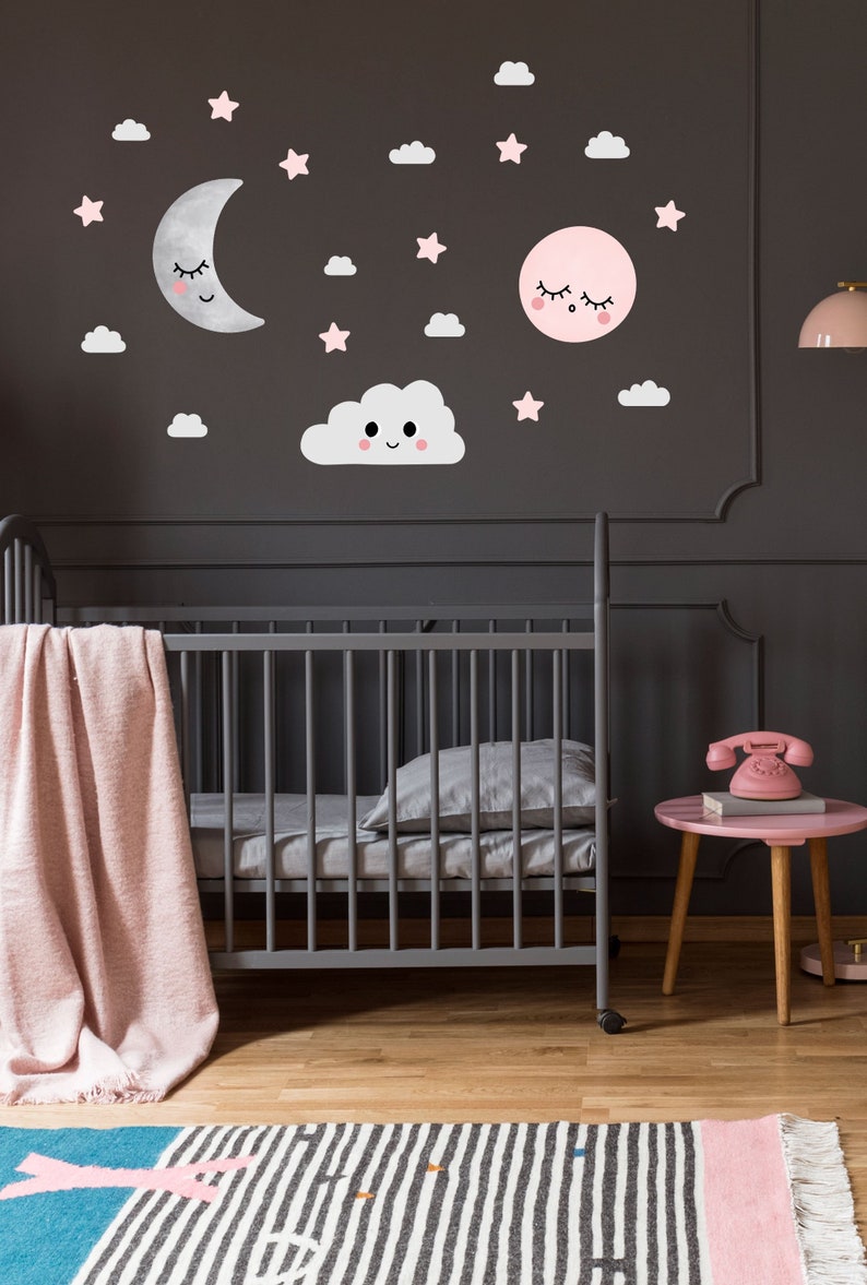 May include: A gray crib with a pink blanket and a gray pillow. The wall behind the crib has pink and white clouds, stars, and moons with smiling faces.