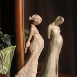 May include: A white ceramic sculpture of a pregnant woman standing with her hands clasped in front of her. The sculpture is on a wooden surface with a stack of books.