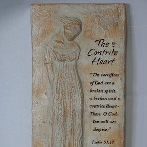 May include: A rectangular, beige-toned wall plaque with a sculpted figure of a person in a long dress. The text "The Contrite Heart" and a Bible verse are printed on the right side. The plaque has a textured surface.