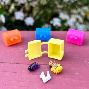 May include: A collection of colorful miniature treasure chests in orange, blue, yellow, and pink. One chest is open, revealing a purple gemstone, a white object, and dried yellow flowers. The chests are arranged on a pink surface.