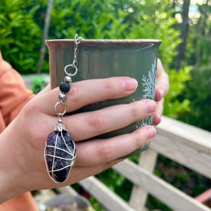 May include: A purple amethyst tea infuser with a silver wire cage and chain, held over a green mug. The infuser features lava rock and silver beads. The mug has a white tree design and a brown rim. The background is blurred green foliage.