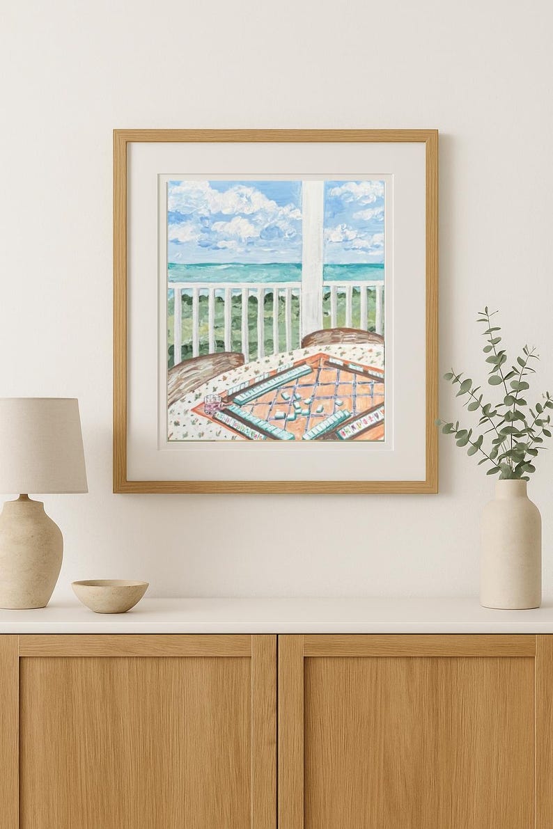 Puede incluir: Una pintura de acuarela enmarcada representa una escena costera con una mesa preparada para un juego. La pintura muestra una vista del oc&eacute;ano y el cielo a trav&eacute;s de una ventana. La mesa tiene un tablero de juego. El marco es marr&oacute;n claro.
