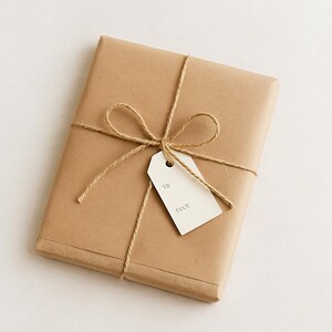 Puede incluir: Un regalo rectangular envuelto en papel marr&oacute;n, atado con hilo y adornado con un lazo. Una etiqueta blanca est&aacute; adjunta con las palabras "TO" y "TRUE" impresas. El regalo se presenta sobre una superficie blanca.
