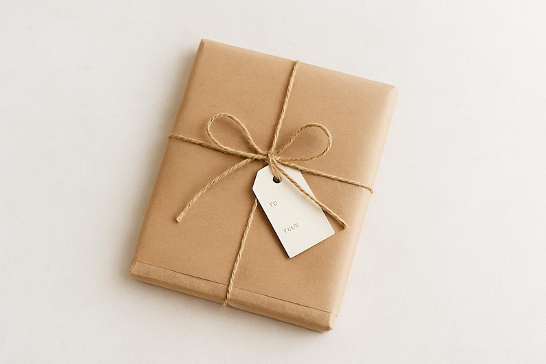 Puede incluir: Un regalo rectangular envuelto en papel marr&oacute;n, atado con hilo y adornado con un lazo. Una etiqueta blanca est&aacute; adjunta con las palabras "TO" y "TRUE" impresas. El regalo se presenta sobre una superficie blanca.