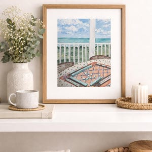 Puede incluir: Una pintura de acuarela enmarcada representa una escena costera con una mesa preparada para un juego. La pintura presenta una vista del mar y el cielo, con una mesa cubierta con un pa&ntilde;o estampado. Un jarr&oacute;n con flores y una taza est&aacute;n en un estante.
