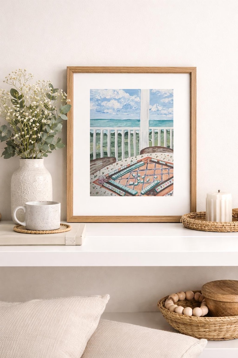 Puede incluir: Una pintura de acuarela enmarcada representa una escena costera con una mesa preparada para un juego. La pintura presenta una vista del mar y el cielo, con una mesa cubierta con un pa&ntilde;o estampado. Un jarr&oacute;n con flores y una taza est&aacute;n en un estante.