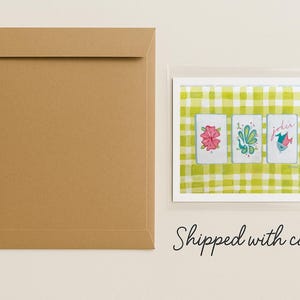 Puede incluir: Un sobre de papel marr&oacute;n junto a una impresi&oacute;n enmarcada de tres naipes. Las cartas presentan ilustraciones de acuarela de flores y un p&aacute;jaro sobre un fondo a cuadros verde y blanco. La palabra "Joker" est&aacute; en una carta.
