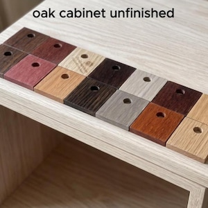 May include: A collection of fourteen small, square wooden samples arranged in two rows, each featuring a single circular hole in the center. The samples display various wood grain patterns and colors, ranging from light tan to dark brown. Text reads: oak cabinet unfinished.