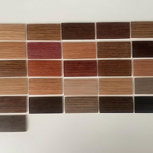 May include: A collection of twenty-six rectangular wood veneer samples arranged in a grid. Each sample displays a unique wood grain pattern and color, ranging from light beige and tan to reddish-brown, dark brown, grey, and a single deep magenta piece.