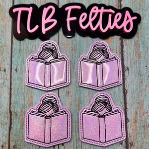 May include: Four embroidered patches featuring a person reading a book, in shades of pink and purple, with black stitching. The patches are on a teal wooden surface. The image also includes a black and pink logo that says "TLB Felties".