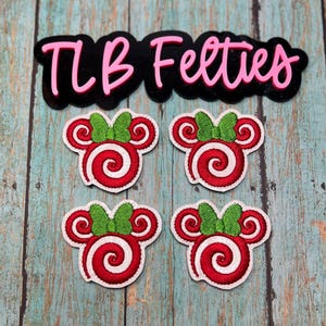 May include: Four red and white candy-themed felt appliques with green bows, arranged below a black and pink text logo that reads "TLB Feeties". The appliques are on a weathered, teal-colored wooden surface.