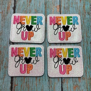 May include: Four white, square patches with a rainbow-colored "Never Grow Up" design and a black Mickey Mouse silhouette.