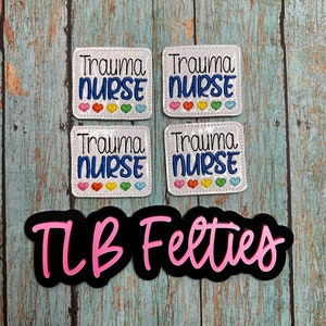 May include: Four white felt patches with the text "Trauma Nurse" in blue embroidery and a row of colorful hearts. Below the patches is a black felt patch with the text "TLB Felties" in pink.