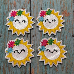 May include: Four embroidered sun patches featuring a white glittery center with a smiling face, surrounded by yellow rays. Each patch is adorned with a small pink, green, and blue flower cluster on the upper left side of the sun's face.