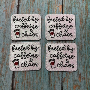May include: Set of four white coasters with a glittery finish. Each coaster features a black and white illustration of a coffee cup with a red heart and the text "fueled by caffeine & chaos".