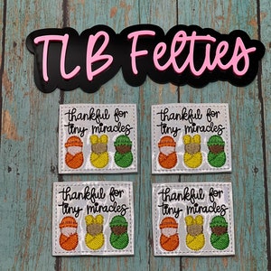 May include: Four white felt patches with embroidered images of three tiny babies in swaddles. Each patch has the text "thankful for tiny miracles" in black cursive.