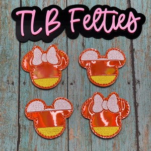 May include: Four felt Mickey Mouse head appliques in orange, yellow, and white. The appliques are shaped like Mickey Mouse heads with a candy corn design. The text "TLB Felties" is visible in the background.