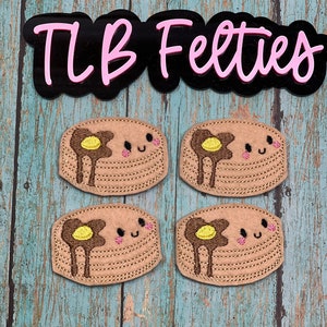 May include: Four brown felt pancake appliques with yellow felt butter and pink felt smiling faces. The appliques are on a blue wooden background with a black sign that says "TLB Felties".