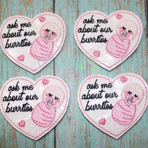 May include: Four heart-shaped embroidered patches with a white glittery background. Each patch features the black text "ask me about our burritos" and an illustration of a baby wrapped in a pink swaddle blanket, accented with small pink hearts.