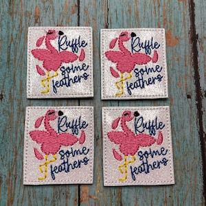 May include: Four white square patches with pink flamingo embroidery. Each patch says "Ruffle some feathers" in blue and yellow thread.