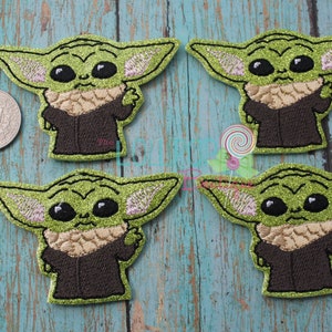 May include: Four green, brown, and pink glitter iron-on patches of a popular fictional character, "The Child" from the Star Wars series. The patches are approximately the size of a US quarter.
