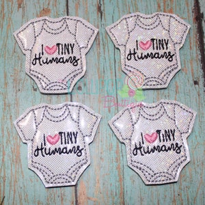 May include: Four white baby onesie-shaped patches with a glittery finish. Each patch features black embroidered stitching details and the text I (heart symbol) TINY Humans in black thread with a pink heart replacing the word love.