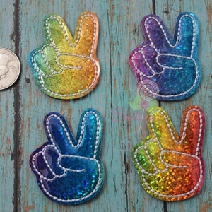 May include: Four glittery peace sign hand patches with white stitched borders. The top left patch is yellow and orange, the top right is purple and blue, the bottom left is blue and teal, and the bottom right is orange and yellow. A US quarter is shown for scale.