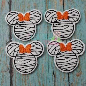 May include: Four white glittery mouse-shaped fabric patches featuring black embroidered mummy-style bandages and a small orange bow between the ears. Each patch has a distinct silhouette with rounded ears and a circular head, showcasing detailed stitching.