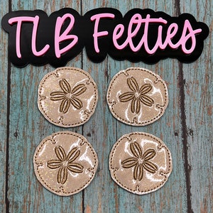 May include: Four beige felt sand dollar coasters with gold embroidery. The coasters are arranged in a square pattern on a blue wooden surface. The text "TLB Felties" is visible in the top center of the image.