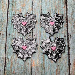 May include: Four black and silver spiderweb patches with a pink heart in the center. The patches are in a web-like shape and are embroidered with white thread. The background is a weathered, blue-green wooden surface.