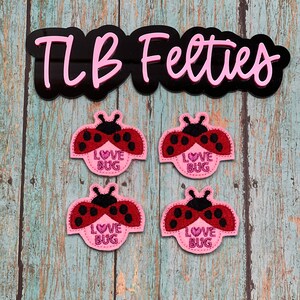 May include: Four red and black ladybug felt appliques with the words "LOVE BUG" on a pink background. The appliques are on a blue and white wooden surface. The image also includes the text "TLB Felties" in pink.
