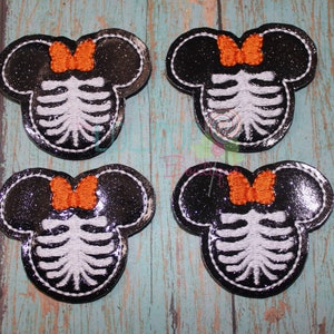 May include: Four black and silver glitter iron-on patches with an orange bow and a white skeleton design. The patches are shaped like Mickey Mouse ears.