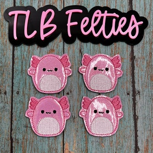 May include: Four pink felt axolotl patches with embroidered details. The patches are on a light blue wooden background. The text "TLB Felties" is visible at the top of the image.