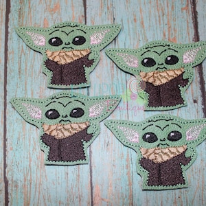 May include: Four embroidered fabric patches featuring a green character with large black eyes, pointed ears, and a brown robe with a textured tan collar. Each patch has a stitched border and displays a consistent design of the same fictional creature.