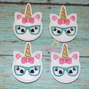 May include: Four embroidered unicorn patches featuring white glittery faces, gold metallic horns, pink bows, and teal glasses. Each unicorn has black embroidered eyes with long eyelashes. The patches are arranged in a two-by-two grid pattern.