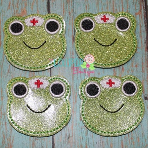May include: Four glittery green frog-shaped patches. Each patch features a smiling frog face with large black eyes, a white nurse's cap with a red cross, and a stitched green outline. The patches are on a teal wood background.