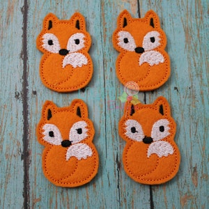 May include: Four orange felt fox appliques with white and black embroidered details. The foxes are sitting with their tails curled around their feet.