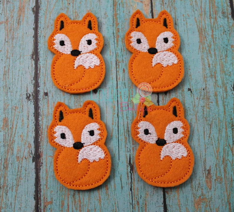 May include: Four orange felt fox appliques with white and black embroidered details. The foxes are sitting with their tails curled around their feet.