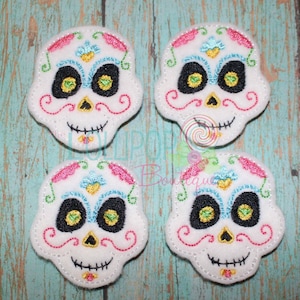 May include: Four white felt sugar skull appliques with colorful embroidery details. The skulls have black eyes, pink and blue accents, and a gold glitter nose.