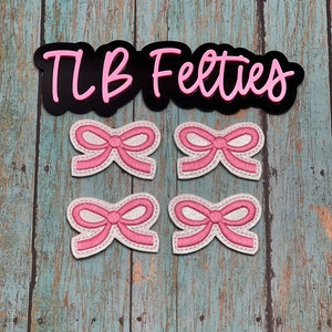 May include: Four pink felt bows with white glitter accents. The bows are arranged in two rows of two. The text "TLB Felties" is visible above the bows.