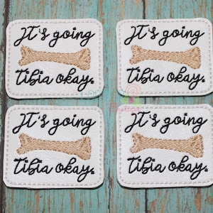May include: Four white square patches with black stitching around the edges. Each patch has a brown bone graphic and the text "It's going Tibia okay." in black cursive.