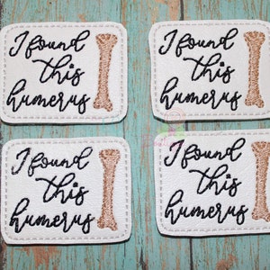 May include: Four white square patches with black stitching around the edges. Each patch has a brown bone image and the text "I found this humerus" in black cursive writing.