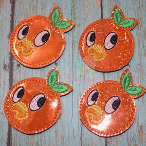May include: Four orange-colored, glittery applique patches shaped like cartoon oranges with green leaves. Each patch has a stitched outline, black eyes, and a yellow beak. The patches are arranged on a blue and teal wooden surface.