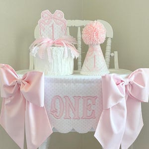 May include: A white high chair decorated for a first birthday. The chair has a pink bow on each side and a banner with the word "ONE". A cake and party hat are on the chair, decorated with pink accents.
