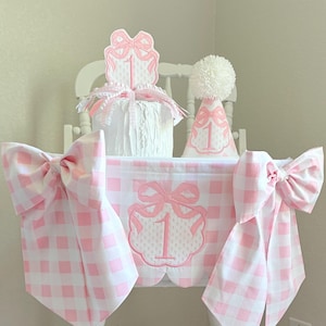 May include: A white wooden high chair decorated for a first birthday. The chair features a pink and white gingham fabric with large bows, a decorative banner with the number "1", and a matching party hat.