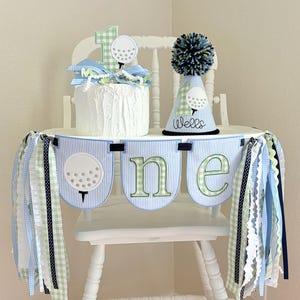 May include: A white high chair decorated for a first birthday. The chair is adorned with a banner spelling "ONE" in green and blue letters, and a cake with a golf-themed topper. A matching party hat sits on the chair.