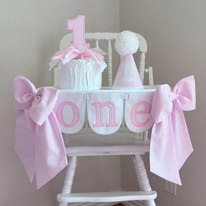May include: A white high chair decorated for a first birthday. It features a pink and white gingham banner that spells "one", a cake with a number "1" topper, and a party hat. Large pink bows adorn the sides.