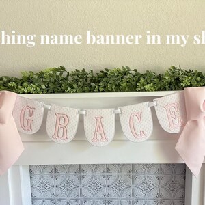 May include: A white banner spelling "GRACE" in pink letters, with a pink bow on each end. The banner is strung across a white shelf with greenery above. The text "Matching name banner in my shop!" is at the top.